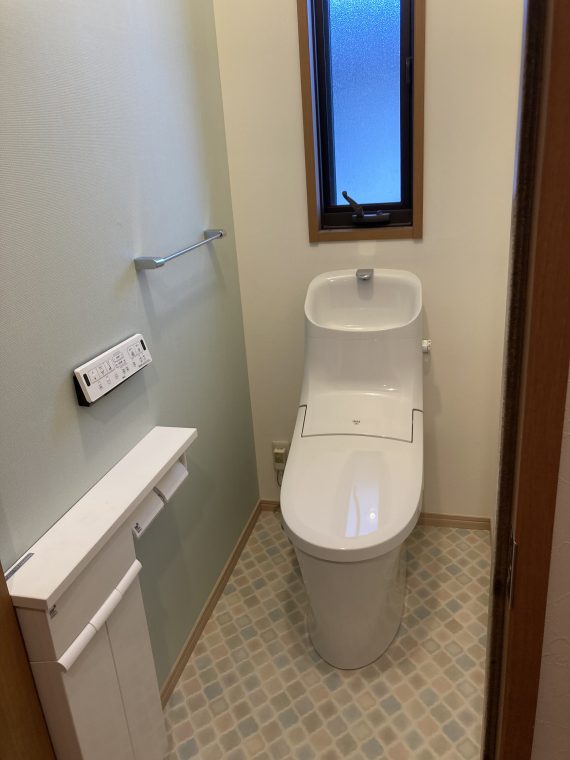 工事後の新しいトイレの写真です。
トイレ便器の背面にタンクがついていて、そこに手洗い水栓がついています。壁は、入って左側が薄緑のクロス、それ以外の三方向は白いクロスです。
床は少しレトロな雰囲気もあるひし形の可愛らしい色合いのクッションフロアです。