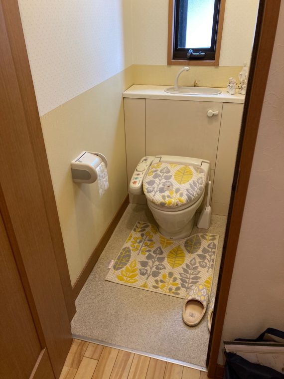 既存のトイレ写真です。
トイレ便器の背面に、手洗いと収納の棚がついています。壁は、下から1メートルの位置まで薄黄色のクロス、1メートルから天井までは白いクロスです。